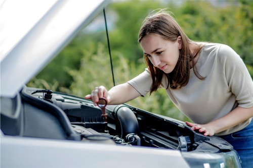Ready to Travel? Don't Skip These 5 Pre-Trip Automotive Services in Kenner, LA. CAMS Automotive. Image of a woman pulls out a probe in her car engine to check the oil level. Oil change service is an important part of pre-trip vehicle inspection.