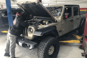 auto repair costs, auto repair in Kenner, LA at CAMS Automotive. Mechanic inspecting the engine of a lifted Jeep Gladiator with the hood open inside an auto repair shop.