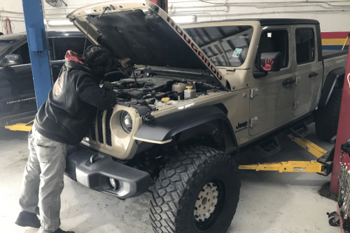 auto repair costs, auto repair in Kenner, LA at CAMS Automotive. Mechanic inspecting the engine of a lifted Jeep Gladiator with the hood open inside an auto repair shop.