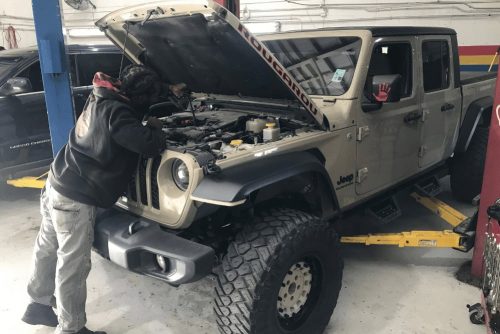 auto repair costs, auto repair in Kenner, LA at CAMS Automotive. Mechanic inspecting the engine of a lifted Jeep Gladiator with the hood open inside an auto repair shop.